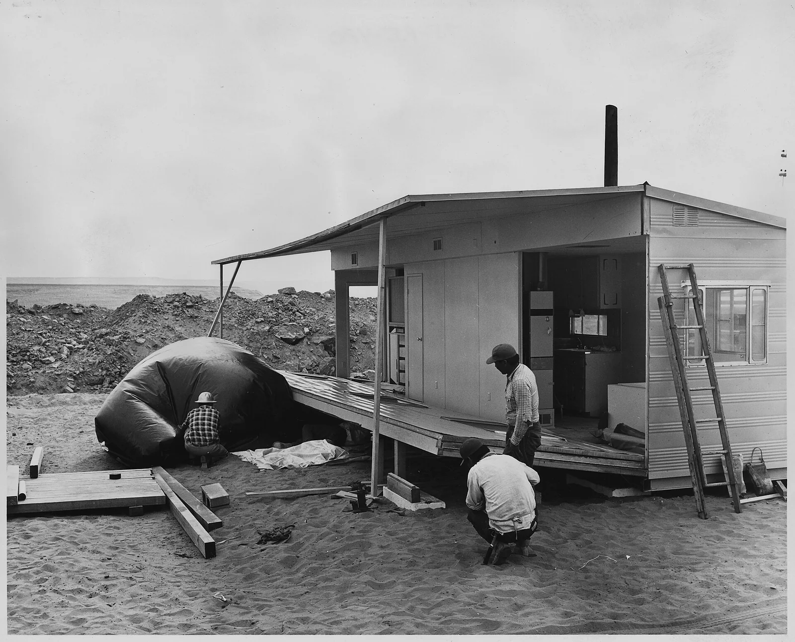 Black-and-white archive photo of a work crew setting up a sectional manufactured home on blocks, with the kitchen visible through the open side