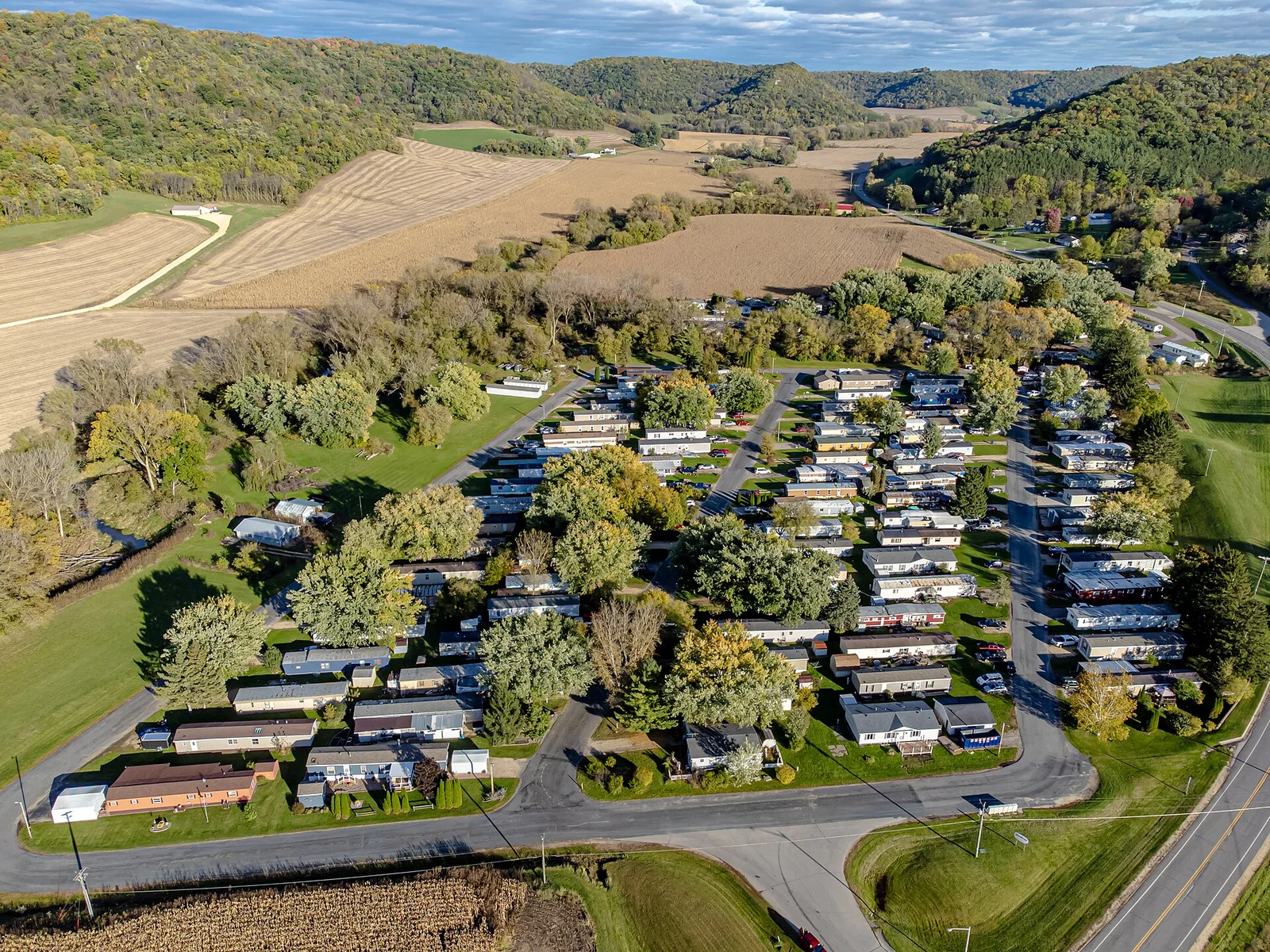 Aerial view of a manufactured home community with rows of single- and double-section homes along paved loops, surrounded by fields and wooded hills